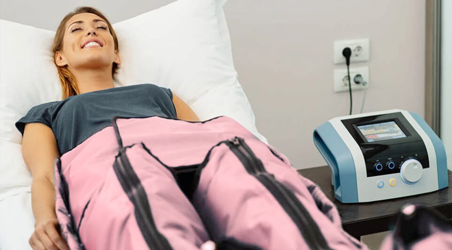 Mujer sonriente y relajada acostada en una camilla, recibiendo un tratamiento de presoterapia con un traje neumático rosa, junto a la máquina de control.